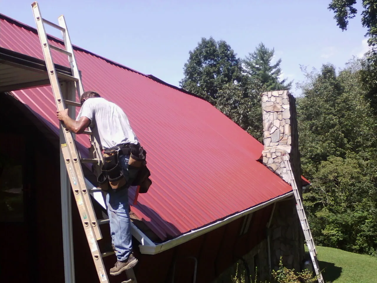Roofer on a ladder beside a red metal roof and stone chimney — Soffit & Fascia Repair by Roofing Brothers in Rio Vista